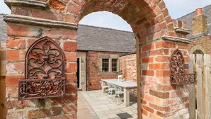 Une entrée en briques en forme d’arc, agrémentée d’éléments décoratifs en métal, qui mène à un patio aménagé avec une table et des chaises.