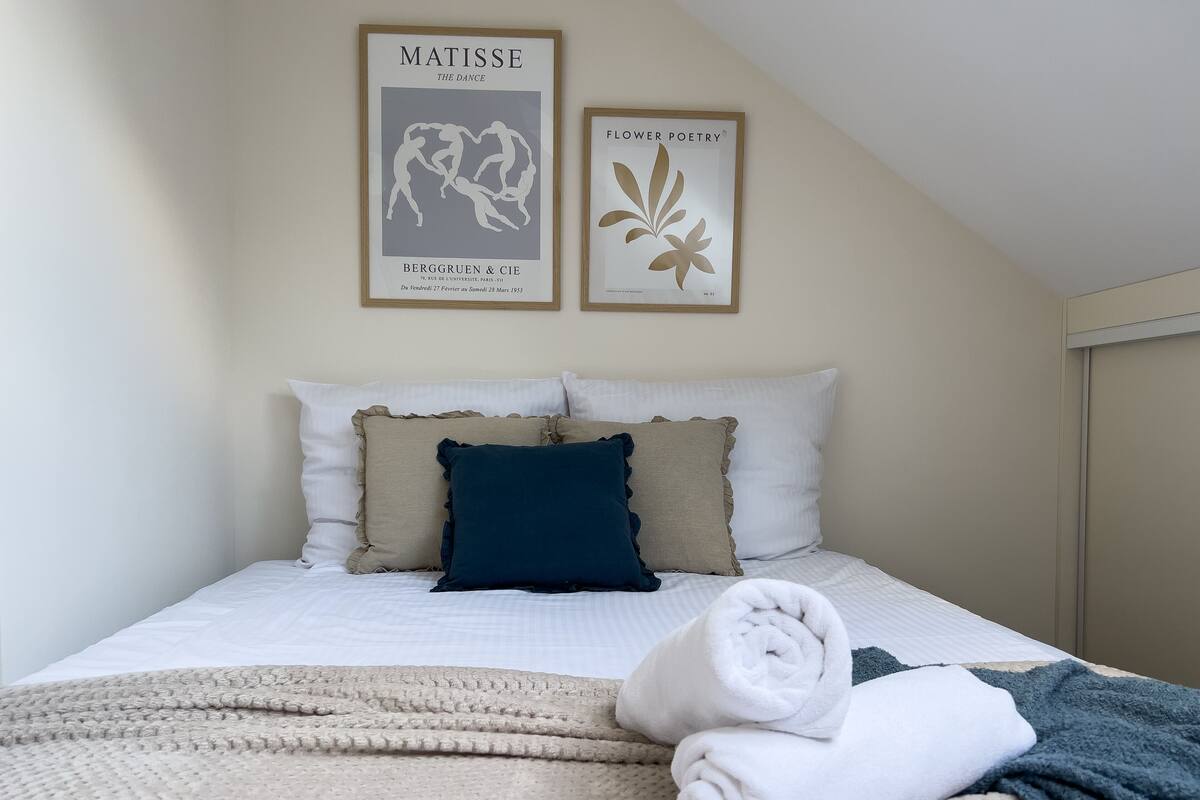 A neatly made bed with white linens, a beige and dark blue pillow, and a beige blanket. There are two framed artworks above the bed: one titled 'Matisse' and another titled 'Flower Poetry'.