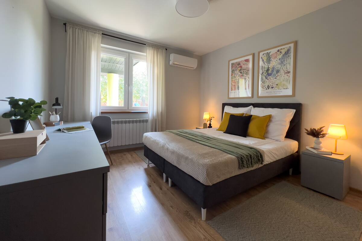 A modern hotel room with a bed, bedside tables, a desk, a chair, a window with curtains, and two framed pictures on the wall.