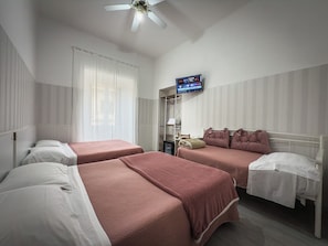 Habitación de hotel con dos camas, ventilador de techo, televisión en la pared y ventana con cortinas traslúcidas.
