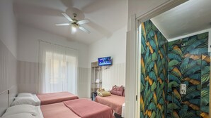 Habitación de hotel con dos camas, ventilador de techo, televisión y zona de ducha con un papel tapiz tropical.