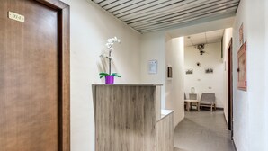 Zona de recepción con un mostrador de madera, una orquídea en maceta y un pasillo que lleva a un área de descanso con sillas.
