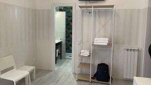 Habitación de hotel con un banco blanco, un armario con estantes, un televisor montado en la pared y una vista parcial de un baño con ducha y toallero.
