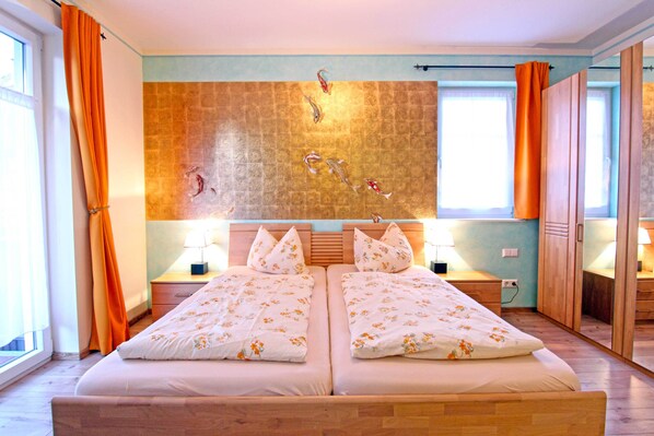 A double bed with floral bedding, two bedside tables with lamps, a wall-mounted artwork, and a view of the outside through a window. - SCHUSTERjunge APARTHOTEL (Sasbachwalden)
