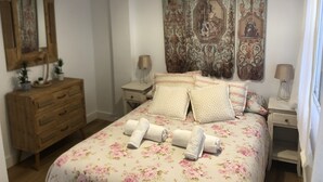 Et soveværelse med blomstret sengetæppe, to sengeborde med lamper, en trækommode og et stort gobelin på væggen.