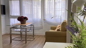 En moderne stue med et glasbord, en beige sofa, en potteplante og et vindue med lette gardiner.