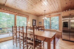 A wooden interior with a dining table, chairs, a refrigerator, and large windows offering a forest view.