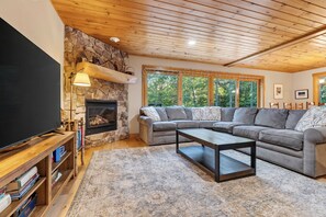 A cozy living room with a stone fireplace, a large flat-screen TV, and a sectional sofa. - Wooded Bethel Chalet | Fireplace, Deck & Fire Pit (Bethel)