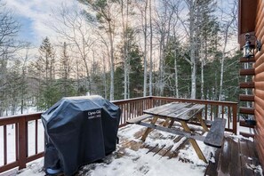 Une terrasse en bois dotée d’un barbecue couvert, d’une table de pique-nique et offrant une vue sur une zone boisée.