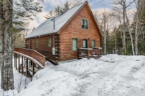 Une cabane en rondins avec un porche et une terrasse, entourée de neige et d’arbres.
