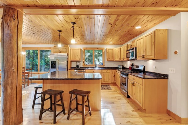 A spacious kitchen with wooden cabinets, a large island with stools, and a view of the forest through the windows. - Wooded Bethel Chalet | Fireplace, Deck & Fire Pit (Bethel)