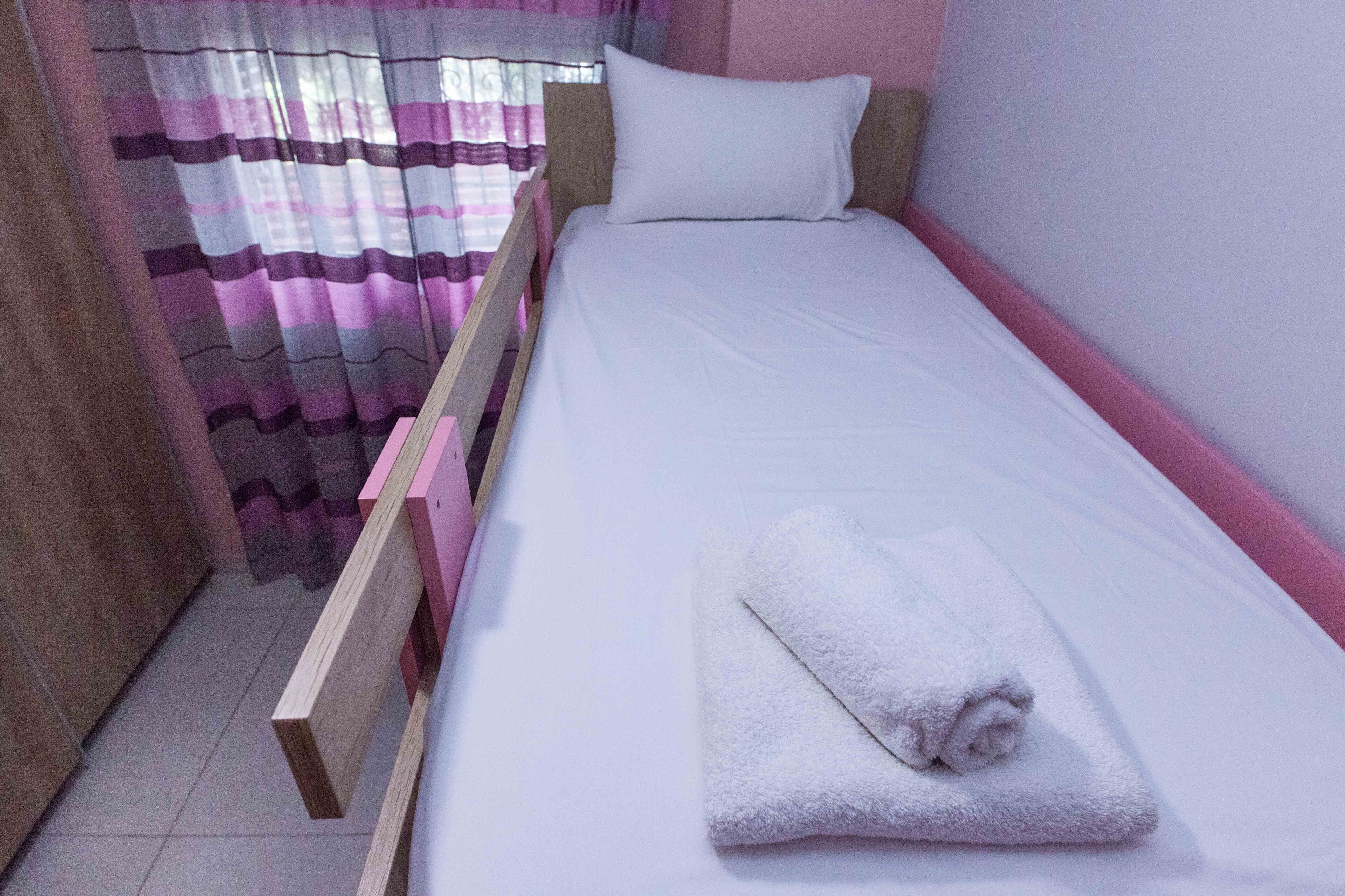 Een enkel bed met een witte matras en een opgerolde witte handdoek, een houten hoofdbord en een roze muur.
