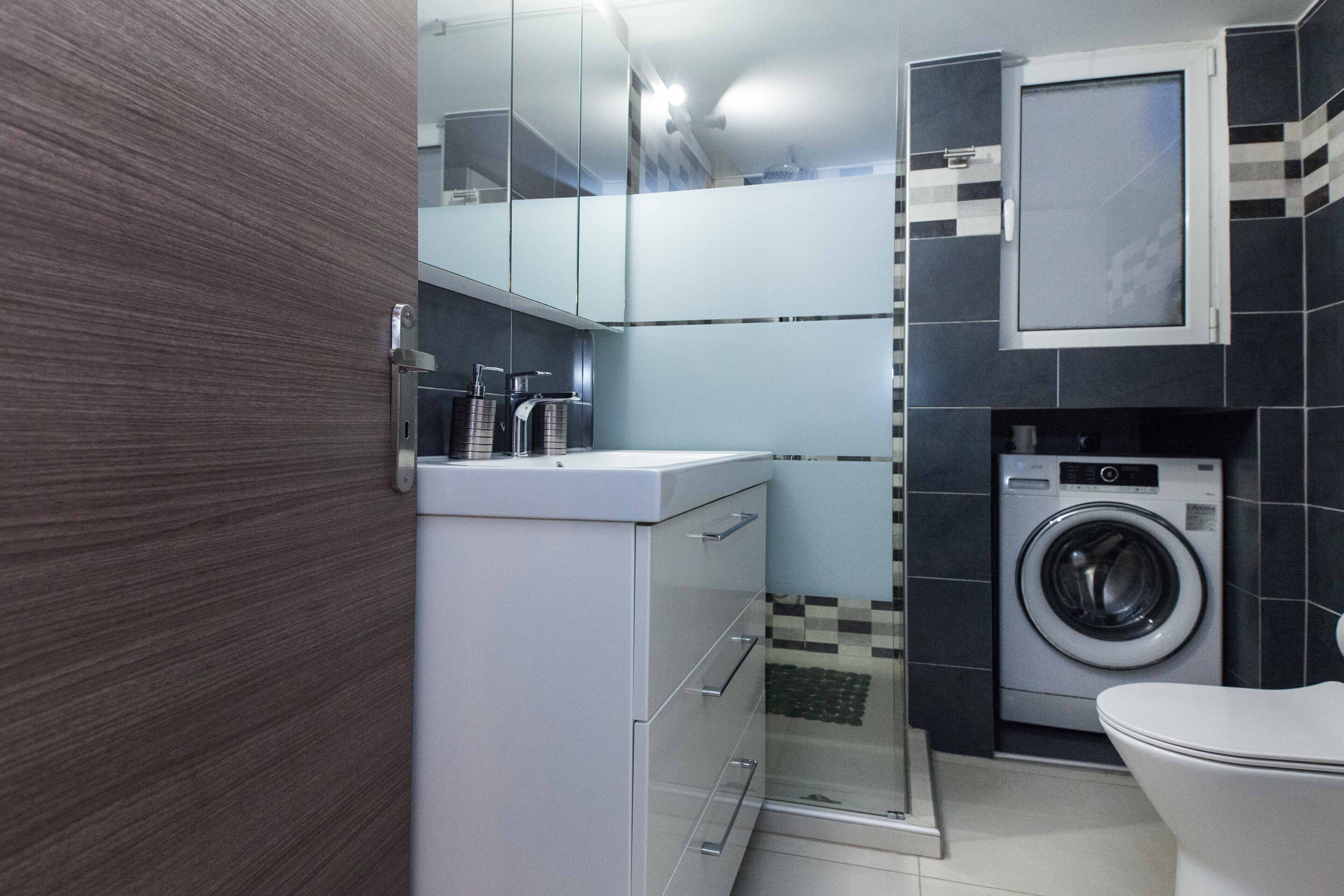 Une salle de bain compacte au design moderne, dotée d’un lave-linge encastré, de toilettes et d’une cabine de douche en verre.