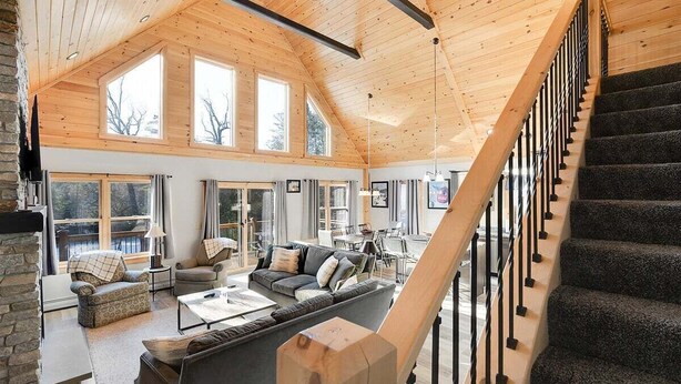 A spacious living area with a high ceiling, large windows, a staircase, and a stone fireplace.