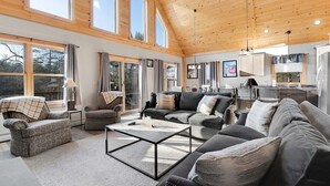 A spacious living room with a high ceiling, large windows, and a kitchen area visible through an open door.