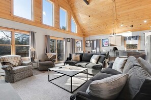 A spacious living room with a high ceiling, large windows, and a kitchen area visible through an open door. - Mountain Lake Retreat- Waterfront Chalet (Woodstock)