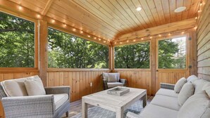 A covered outdoor seating area with wicker furniture, a wooden table, and large windows.