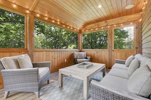 A covered outdoor seating area with wicker furniture, a wooden table, and large windows. - Mountain Lake Retreat- Waterfront Chalet (Woodstock)