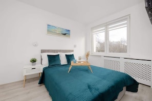 A bedroom with a bed, a bedside table with a plant, a window with blinds, and a radiator. - Functional Apartment in the Center | Close to the Sea and Balcony (Gdynia)