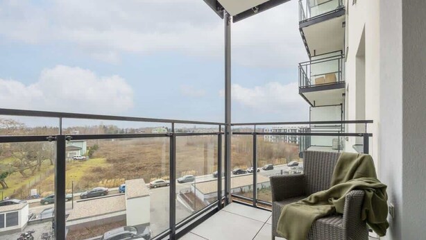A balcony with a wicker chair and a blanket, overlooking a parking area and buildings. - Charming Apartment with Balcony | Close to Chopin Airport (Warszawa)
