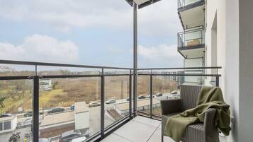 A balcony with a wicker chair and a blanket, overlooking a parking area and buildings.