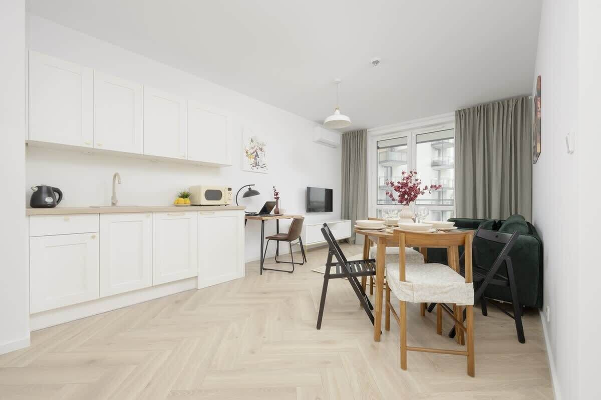 A modern kitchen with white cabinets, a dining area with a wooden table and chairs, and a large window with curtains.