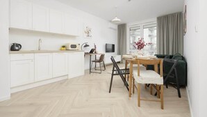 A modern kitchen with white cabinets, a dining area with a wooden table and chairs, and a large window with curtains. - Charming Apartment with Balcony | Close to Chopin Airport (Warszawa)