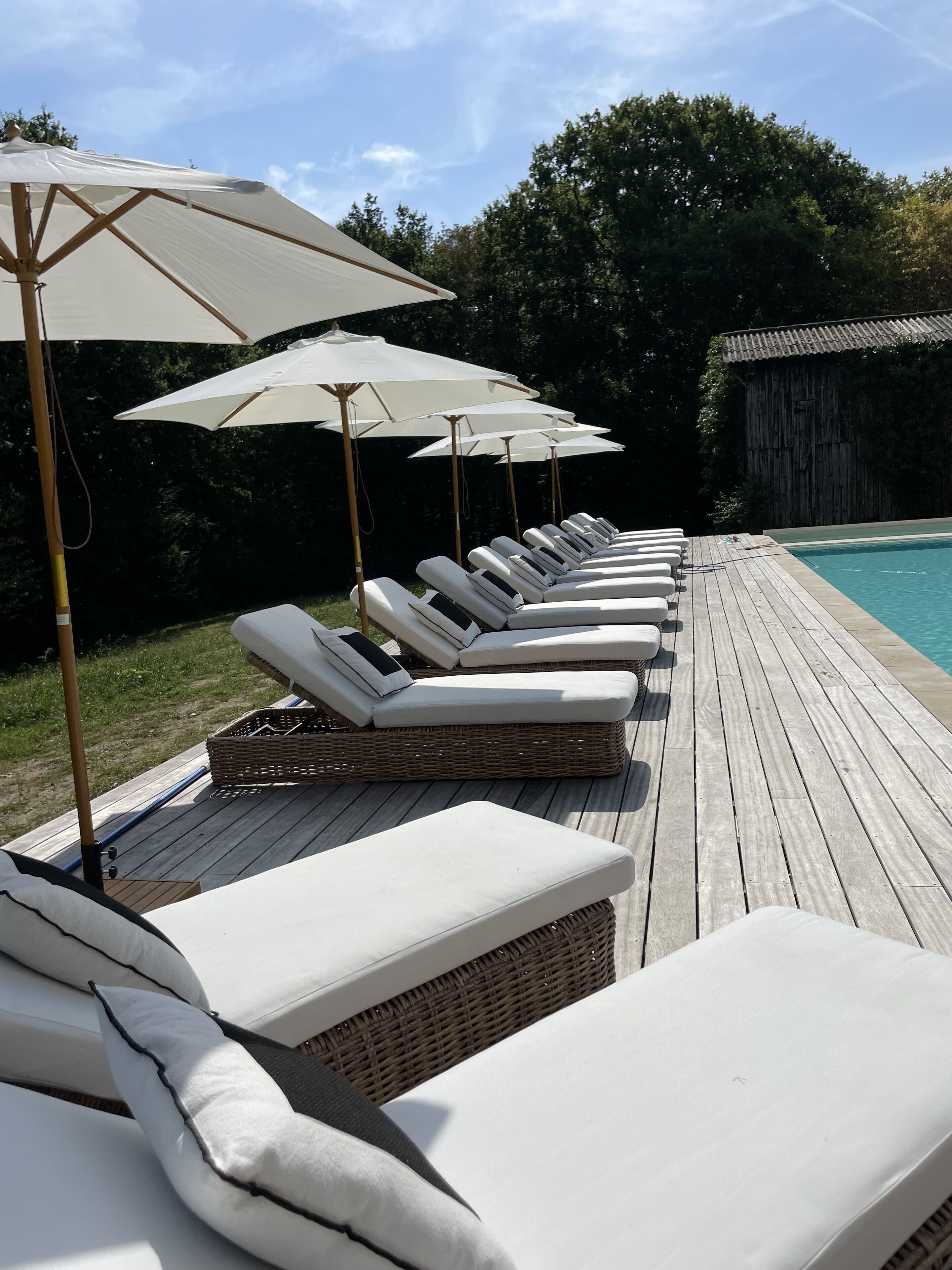 A row of sun loungers with white cushions and beige umbrellas on a wooden deck by a pool.