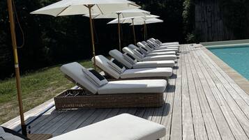 A row of sun loungers with white cushions and beige umbrellas on a wooden deck by a pool.