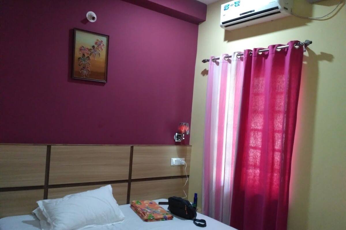 Un dormitorio con cama, mesita de noche, calefactor y ventana con cortinas rosas.