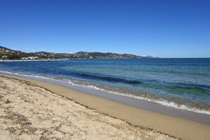 A beach with clear blue water, sandy shore, and distant hills.