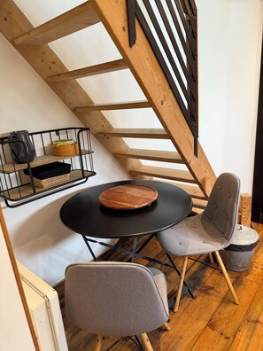 A cozy dining area with a round table, two chairs, a wooden staircase, and a wall-mounted shelf with baskets. - A natural, cozy oasis just 25 minutes from Bordeaux. (LA LANDE DE FRONSAC)
