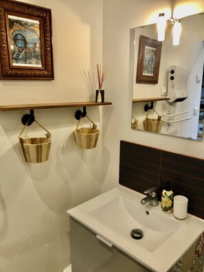 Un baño con un lavamanos blanco, un espejo, un estante de madera con objetos decorativos y un cuadro en la pared.