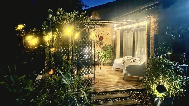 A covered outdoor seating area with a table and chairs, surrounded by plants and string lights.