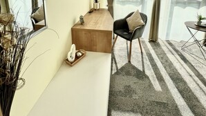 A hotel room with a large bed, a desk with a flat-screen TV, a chair, and a view of the sea through large windows. - LikeDaheim Apartments (Rosbach vor der Hoehe)