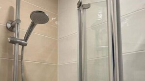 Une douche équipée d’un pommeau de douche à main et d’un robinet mural, d’une paroi de douche en verre et d’une petite étagère blanche où sont rangés les articles de toilette.