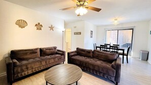 A living room with two brown sofas, a round coffee table, a ceiling fan, and a dining area with a table and chairs.