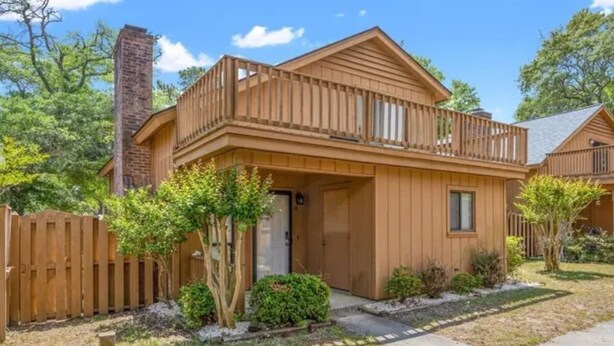 A two-story wooden house with a balcony, a brick chimney, and a small front yard with trees and shrubs.