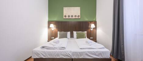 Uma cama de casal com estrutura de madeira, duas mesas de cabeceira com abajures, uma parede verde e uma janela com cortinas.