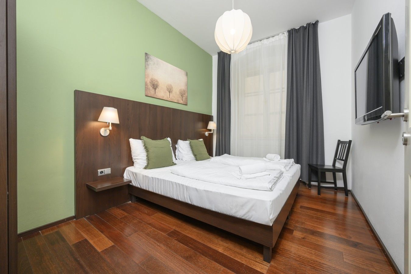 Une chambre d’hôtel avec un lit en bois, deux lampes de chevet, un tableau au mur, un téléviseur fixé au mur et une chaise.