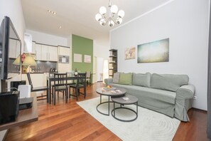 A modern living room with a grey sofa, a dining table with chairs, a TV, and a kitchen area in the background.