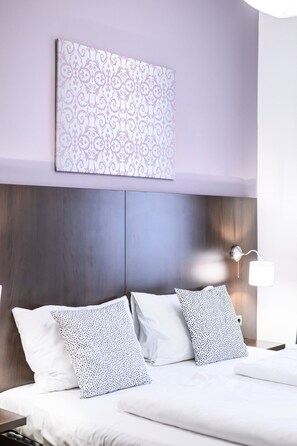 Quarto de hotel com cama, duas almofadas, luminária fixada na parede e um objeto decorativo na parede.