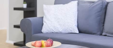Un salon moderne avec un canapé gris, une table basse ronde sur laquelle se trouve un saladier rempli de fruits et quelques magazines, et une bibliothèque noire adossée au mur.