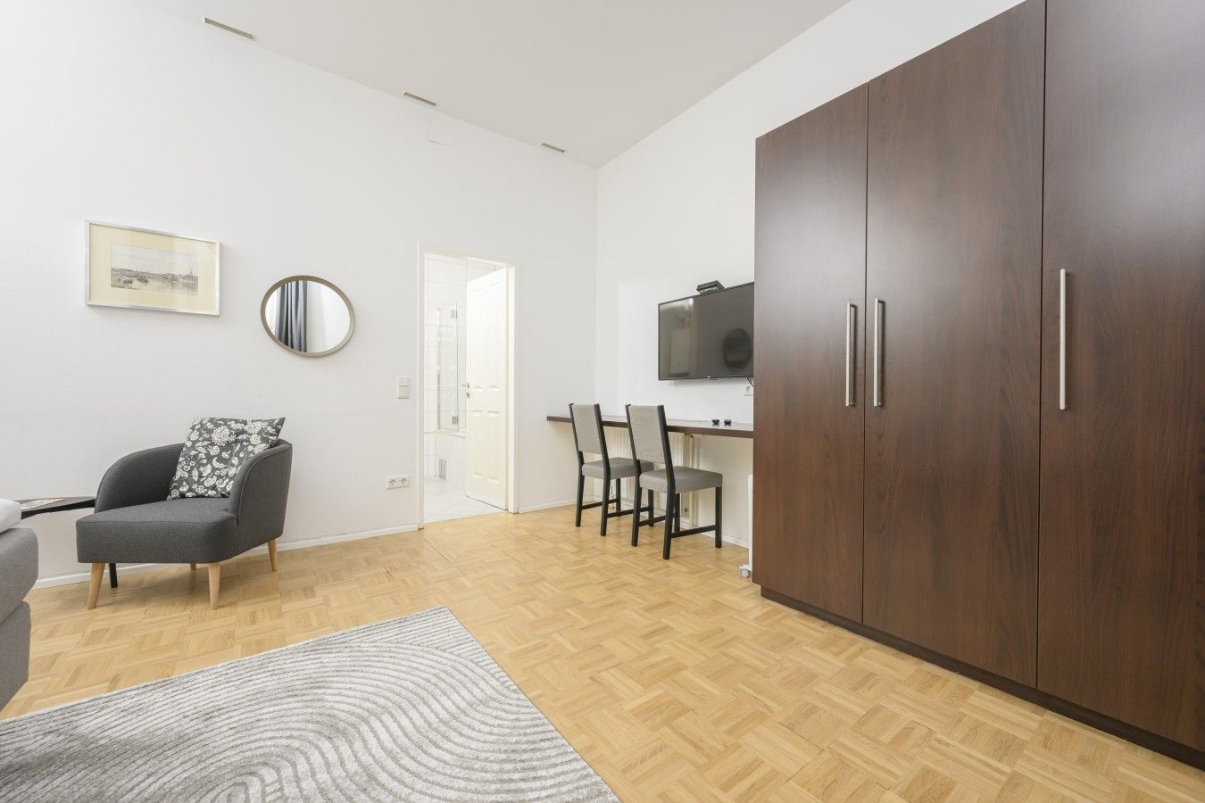 Ein modernes Zimmer mit Holzboden, einem grauen Sessel, einem Esstisch mit Stühlen, einem Fernseher und einem Kleiderschrank.