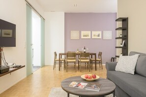 Una sala de estar con un sofá, una mesa de centro con revistas, una mesa de comedor con sillas y un televisor en la pared.