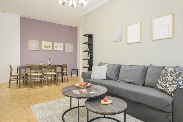 A modern living room with a grey sofa, a dining table with chairs, and a coffee table with magazines and apples.