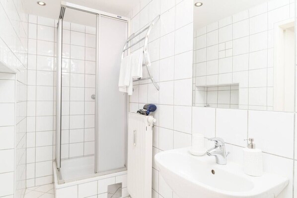 Un baño moderno con ducha envuelta en vidrio, azulejos blancos, un lavamanos blanco con espejo y un perchero para toallas.