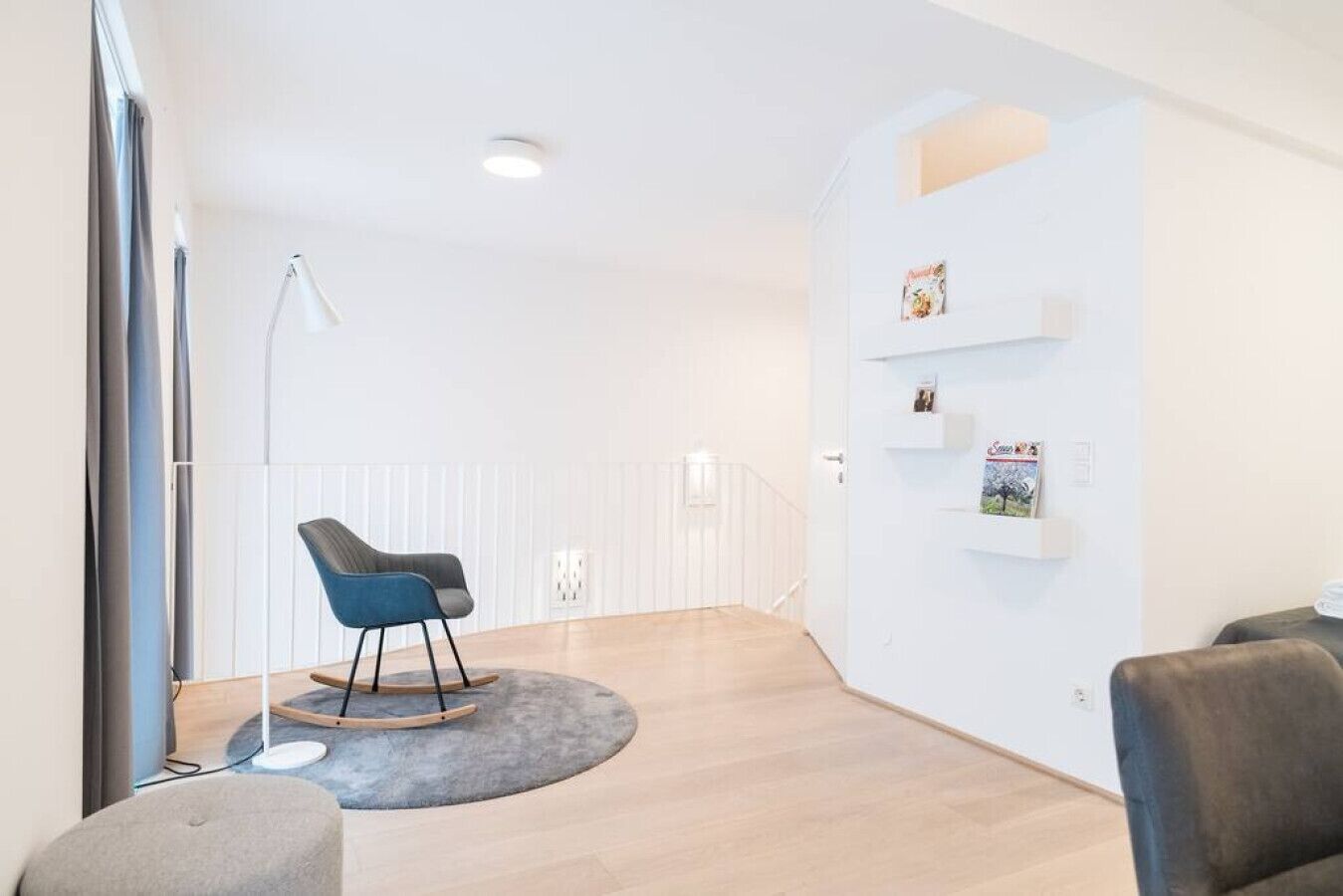 A modern, minimalist room with a blue rocking chair, a grey ottoman, and a white shelving unit with books.