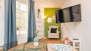 A cozy living room with a green accent wall, a flat-screen TV, a comfortable armchair, a coffee table, and a dining table.