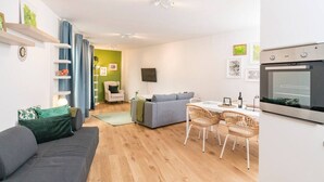A modern living room with a sofa, a dining table with chairs, a TV mounted on the wall, and a built-in oven.